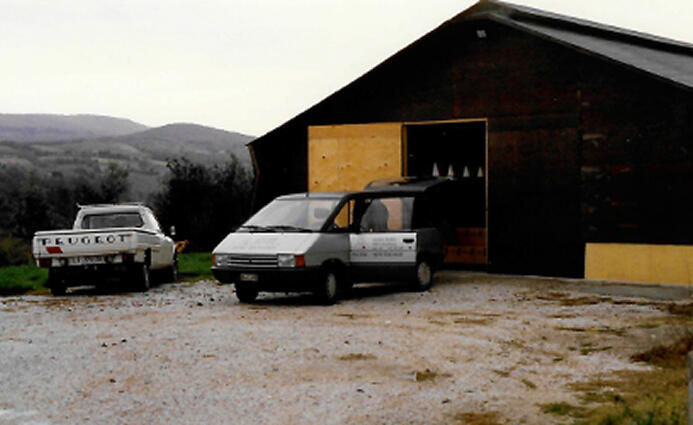 Außenansicht einer einfachen Lagerhalle mit offener Holztür. Davor parken zwei Fahrzeuge, ein Peugeot-Pick-up und ein Lieferwagen.