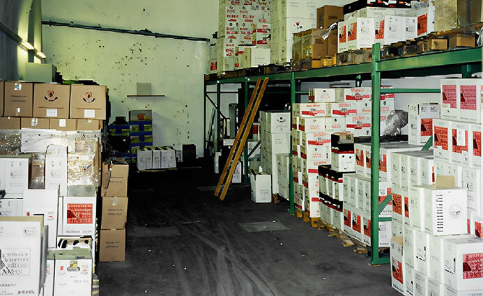 Innenansicht eines Lagers mit gestapelten Weinkartons auf Paletten und Regalen, dazu eine Leiter für die oberen Lagerflächen.
